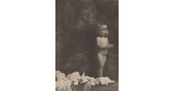 A sepia-toned photograph of a nude White toddler standing next to what looks like a pile of light-colored clothes.