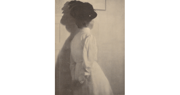 A black-and-white photograph of the back of a White woman wearing a black hat with a feather and a long-sleeved white dress, hands clasped behind her back.