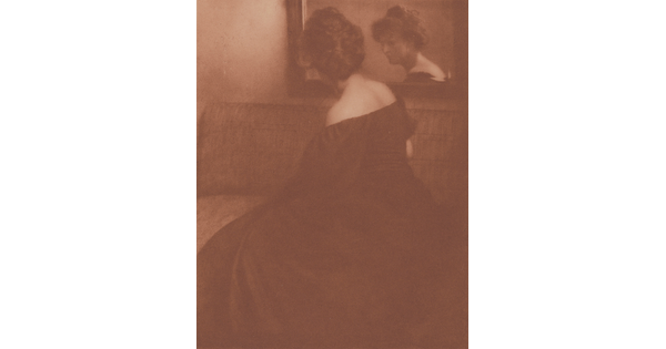 A sepia-toned photograph of a seated White woman, viewed from the side, wearing a long off-the-shoulder dress; her face is visible in the mirror she looks into.