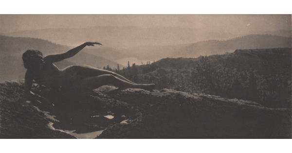 A sepia-toned photograph of a nude woman in shadow lying on a hilltop with cascading hills behind her.