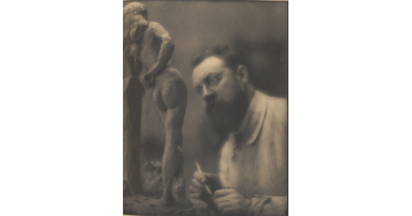 A sepia-toned photograph of a White man with glasses and a beard, holding a sculpting tool in his hand, looking a small sculpture of a nude woman.