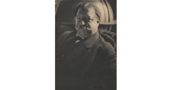 A sepia-toned portrait photograph of a White man with a curled mustache wearing a suit jacket and tie.