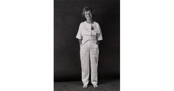 A black-and-white portrait photograph of a standing older White woman with short curly hair, hands in her pockets, wearing a white top, pants and sandals.