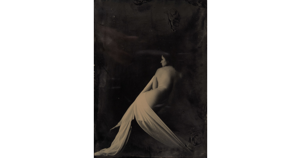 A black-and-white photograph of a seated nude White woman lightly draped in fabric with her back to the camera.