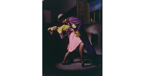 A color photograph of a White man and woman dancing in a dark room in formal attire while a Black child watches through a window.