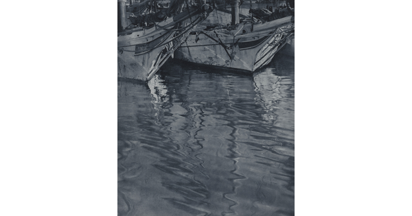 A black-and-white photograph of the ends of two docked sailboats floating on rippling water.
