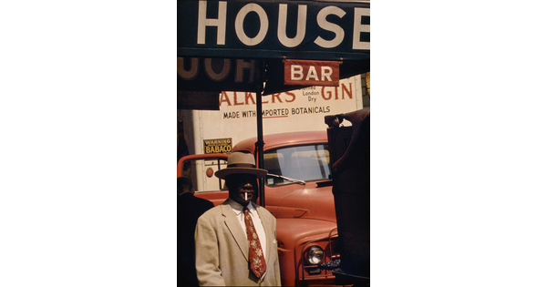 A color photograph of a Black man in a beige hat and suit with a cigarette in his mouth in front of a red car and under a sign that says, "House Bar."