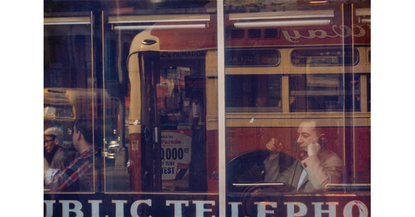 A color photograph of a White man sitting at a cafe window speaking on a phone, with reflections of a passing bus in the glass.