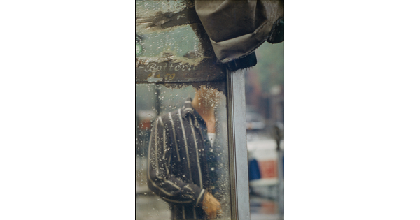 A color photograph of a man's torso through the rain-splatted glass of what looks like a bus shelter.