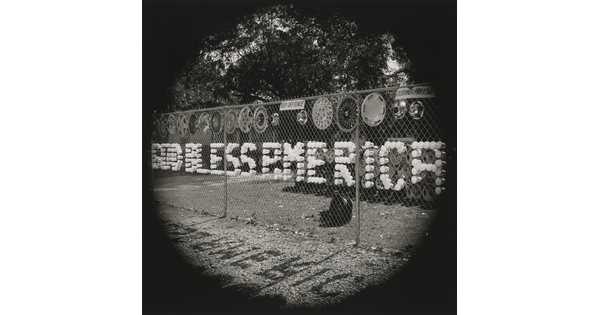A black-and-white photograph of plastic cups arranged in a chain-link fence to spell out "God Bless America."