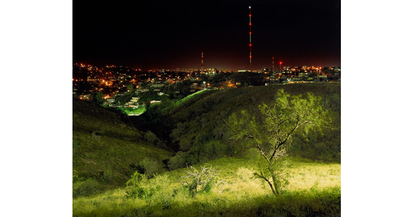 A color photograph of an brightly lit green landscape at night with city lights in the background.