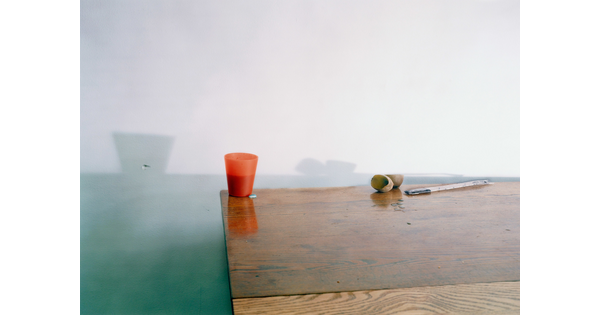 A color still life photograph of a plastic orange cup, a piece of fruit cut in half, and a flat piece of metal on a linoleum table casting shadows against a wall.