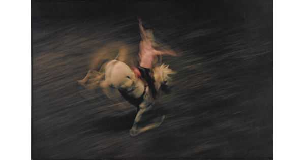 A color photograph of a cowboy on a bucking white horse, with the horse and cowboy blurred, indicating motion.