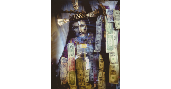 A color photograph of a sculpture of Jesus Christ wearing a crown of thorns behind glass that has strips of dollar bills hanging on it.