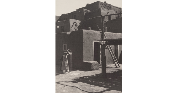 A black-and-white photograph of a woman pouring the contents of a bowl to the ground in front of a multi-level adobe pueblo.
