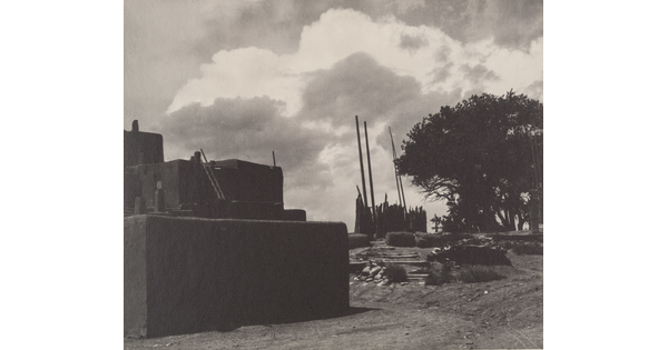 A black-and-white photograph of multi-level adobe pueblo with tall, wood poles standing upright in the distance under a cloudy sky.