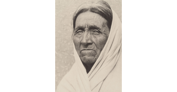 A black-and-white portrait photograph of an elderly Indigenous man with deep wrinkles on his face, a white cloth or shawl covering his head and shoulders.