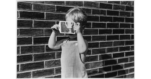 A black-and-white photograph of a White toddler in overalls holding in front of his face an upside down photo of himself standing in front of a brick wall as he stands in front of a brick wall.