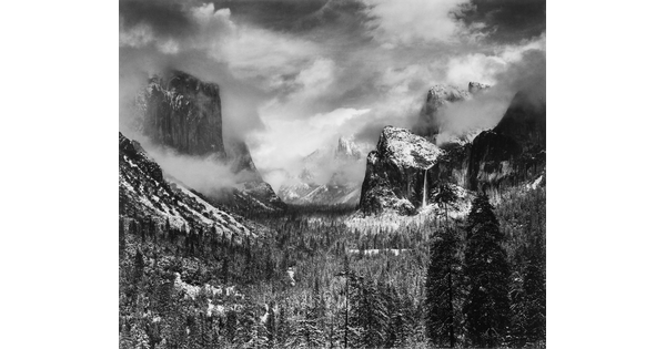 A black-and-white landscape photograph of trees and snowy mountains partially covered in clouds and fog.