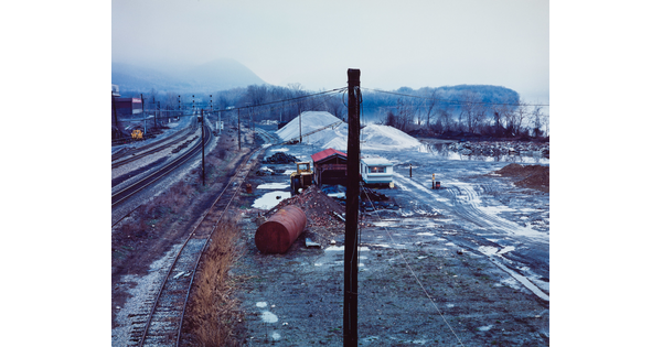 A color photograph of a railyard in winter with rusty barrels, piles of sand, machinery, and patches of snow scattered around with trees and a foggy mountain in the distance.