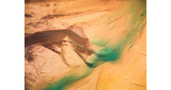 A color photograph of sand dunes from the air with a bright blue-green streak across them.