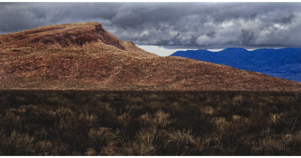 A color photograph of a desert landscape with a dry, brown hill in the foreground and blue mountains in the background under a sky of gray clouds.