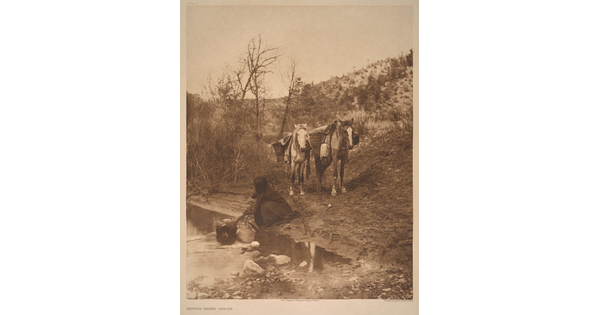 A sepia-toned photograph of an Indigenous person kneeling next to a stream to fill a jug with water as two packhorses wait in the background.