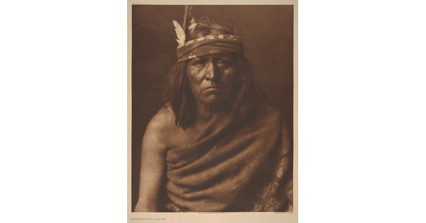 A sepia-toned portrait photograph of an Indigenous man with medium-length hair and a cloth around his forehead and cloth wrapped over one shoulder and around his torso.