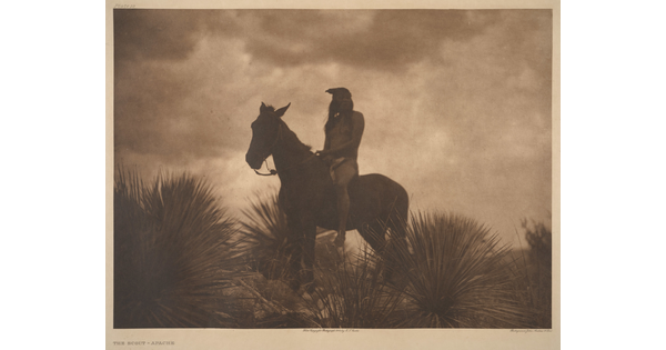 A sepia-toned photograph of an Indigenous person sitting atop a horse surrounded by yuccas; the low light makes the figures look almost like silhouettes.