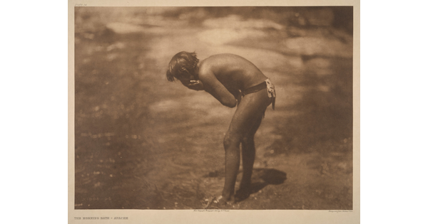 A sepia-toned photograph of a young Indigenous person leaning over a riverbank washing their face.