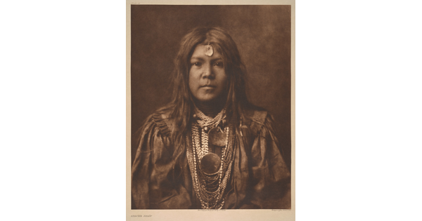 A sepia-toned portrait photograph of a young Indigenous person with long hair, a piece of jewelry on their forehead, multiple stings of beads around their neck, and wearing cultural dress.