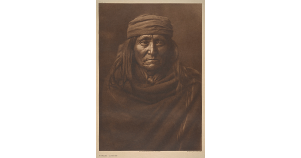 A sepia-toned portrait photograph of an older Indigenous person with long hair, wearing a cap, and wrapped in a solid-colored cloth.