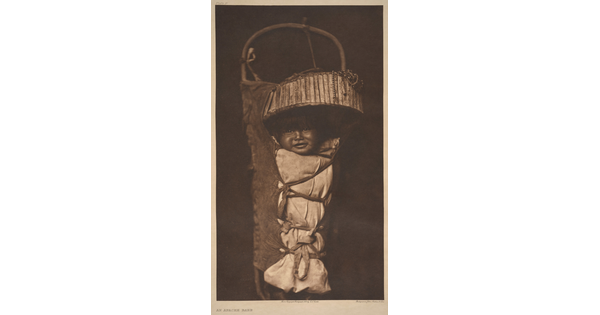 A sepia-toned photograph of a smiling Indigenous baby who is strapped into a traditional cradleboard.