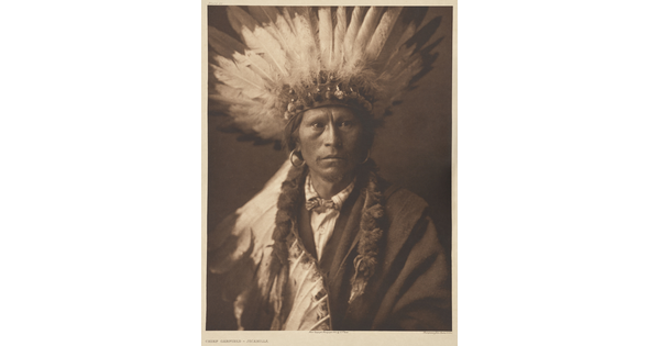 A sepia-toned photograph of a Native American man posed in a suit and feathered headdress.