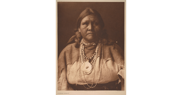 A sepia-toned portrait photograph of an older Indigenous woman with her hair bound on both sides of her head and wearing several beaded necklaces and traditional clothing.