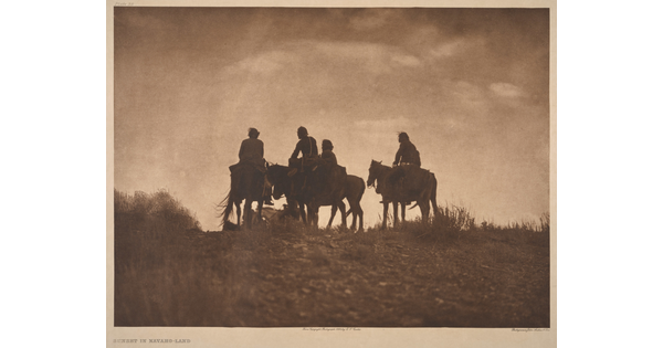 A sepia-toned photograph of the silhouettes of four people on horseback at the top of a hill.