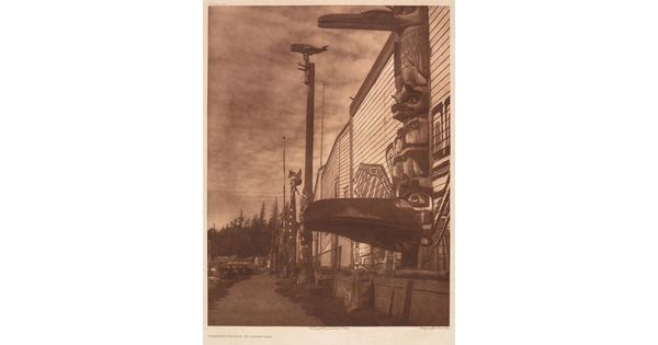 A sepia-toned photograph of several totem poles pressed next to a building with a mural painted on the side.