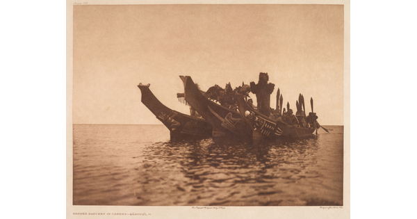 A sepia-toned photograph of three elaborately carved canoes side-by-side on a body of water with several masked Indigenous people in each canoe.