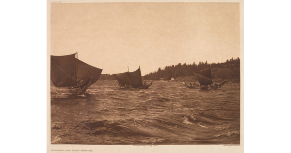 A sepia-toned photograph of three elaborately carved canoes with raised sails on choppy water.