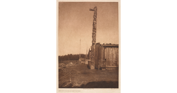 A sepia-toned photograph of a tall totem pole next to a wooden building with a forest in the background.