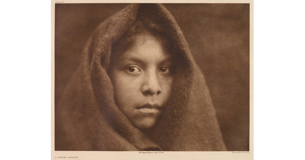 A sepia-toned portrait photograph of a young Indigenous girl wearing a cloth over her head, exposing only her face and hair above her forehead.