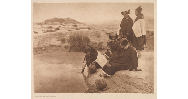 A sepia-toned photograph of a group of five Indigenous women wearing traditional dress sitting and standing on the edge of a spring.