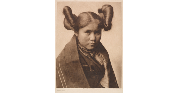 A sepia-toned portrait photograph of a young Indigenous woman with her hair split into two large buns on both sides of her head wearing traditional clothes and necklaces.