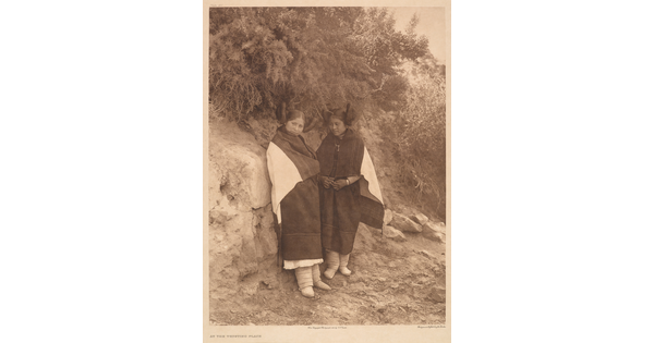A sepia-toned photograph of two Indigenous women in traditional clothing leaning on a rock that has vegetation growing off the top of it.