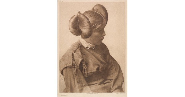A sepia-toned portrait photograph of a young, Indigenous girl wearing a cultural hairstyle and a long sleeved dress.