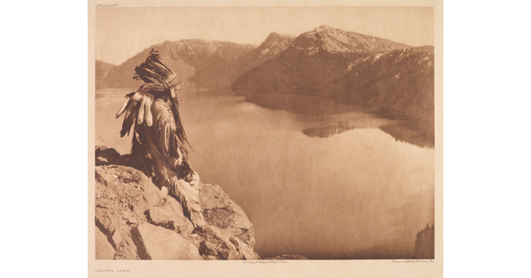 A sepia-toned photograph of an Indigenous man in traditional clothing and a feathered headdress sitting on a rock high above a lake at the base of mountains.
