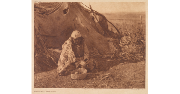 A sepia-toned photograph of an Indigenous person in traditional dress sitting on the ground outside of a hut with a woven basket in front of them.