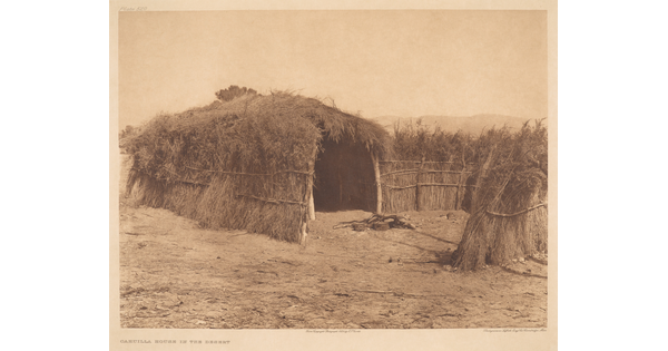 A sepia-toned photograph of a hut made of grass, brush, and wood on a dirt clearing.