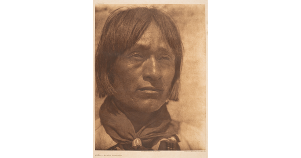 A close-up, sepia-toned portrait photograph of an Indigenous man with hair to his jawline, wearing a scarf around his neck held by a silver concho-style scarf slide.