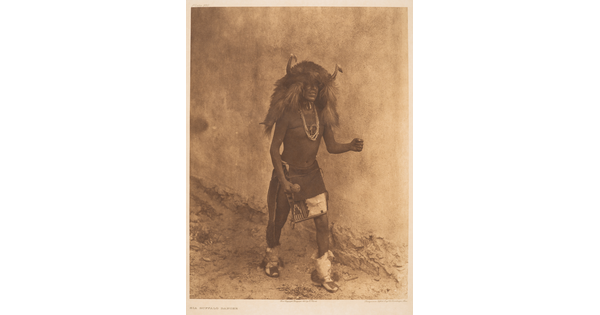 A sepia-toned photograph of an Indigenous man in cultural dress that includes a headpiece made of buffalo hide and horns.